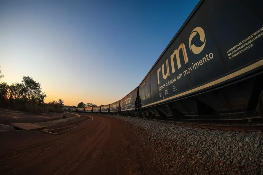 Trem de carga da Rumo Logística circulando ao entardecer, com a frase “Somos o Brasil em movimento” estampada no vagão.
