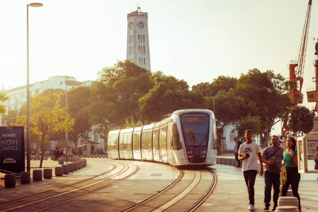 VLT branco circulando em uma praça urbana com pedestres durante o pôr do sol. A mobilidade urbana e o transporte de passageiros também se beneficiam do marketing de conteúdo para empresas de transportes.