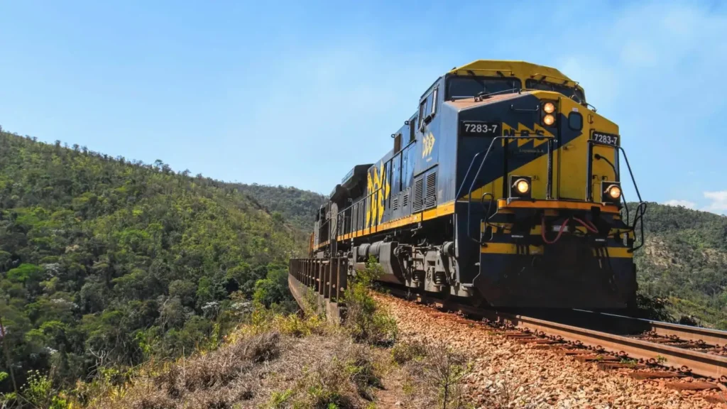 Locomotiva amarela e azul da MRS Logística em movimento sobre trilhos, cercada por montanhas e densa vegetação verde. A ferrovia é um modal estratégico no marketing de conteúdo para empresas de transportes.