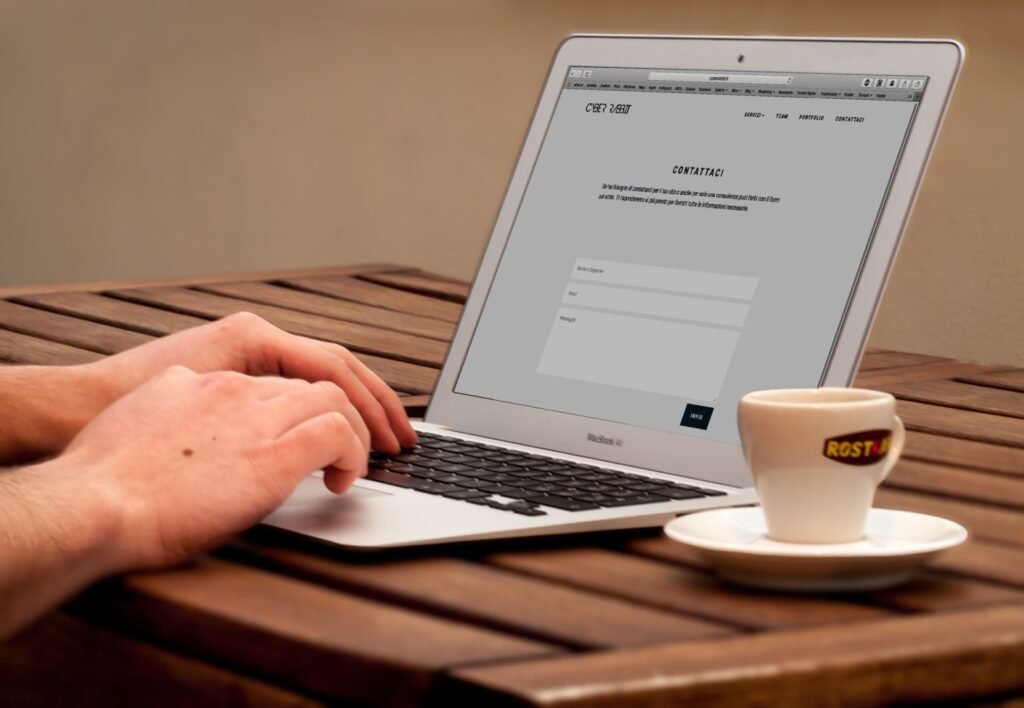 Close-up de mãos digitando em um laptop ao lado de uma xícara de café em uma mesa de madeira.