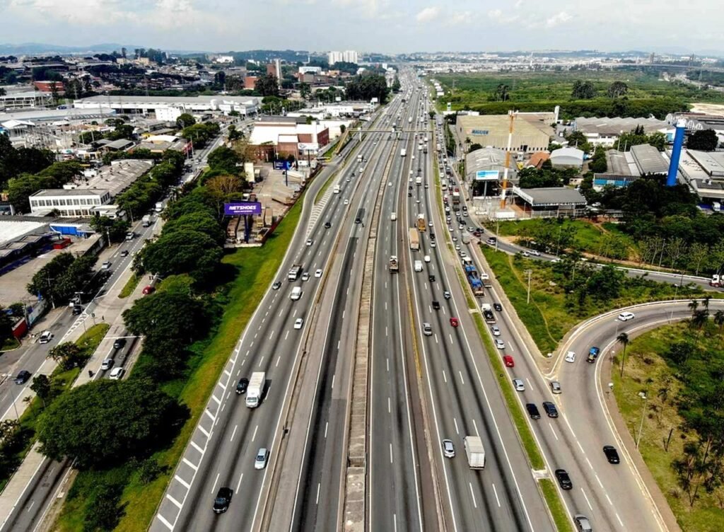 Vista aérea de rodovia com múltiplas pistas e tráfego intenso de caminhões e carros entre áreas industriais